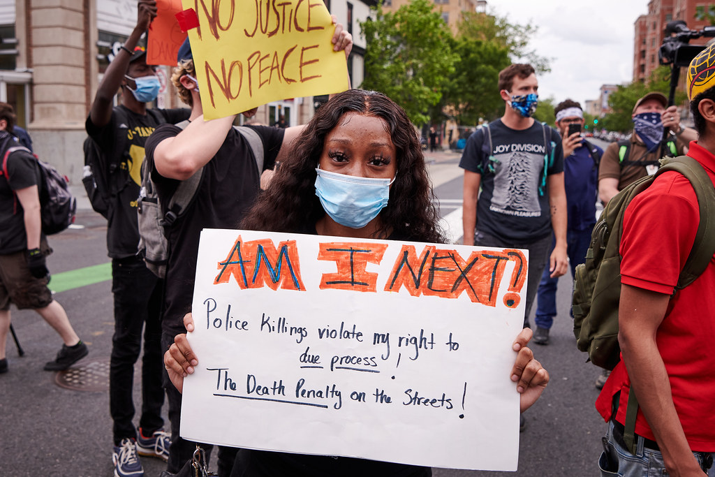 A young Black woman with long, wavy hair parted in the middle, stands at a protest, wearing a face mask and holding a sign that says: "AM I NEXT? Police killings violate my right to due process! The Death Penalty on the streets!"
