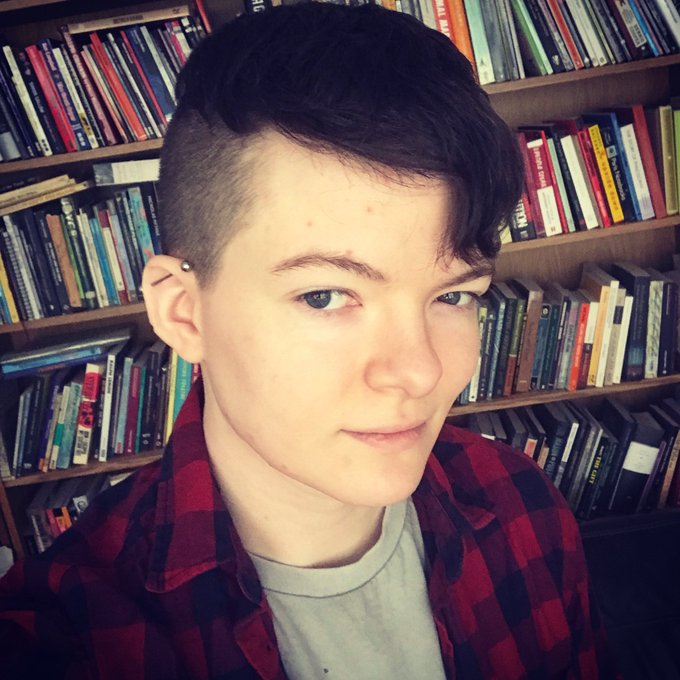 Griffyn is angled away from the camera, but looks back at us with an eyebrow slightly raised and a slight, mischievious smile. His dark hair is buzzed on the sides and longer on top. He has pale skin and wide, dark eyes. He wears a grey T-shirt with a red flannel checked shirt over it and sits in front of a bookshelf stuffed full of books. 