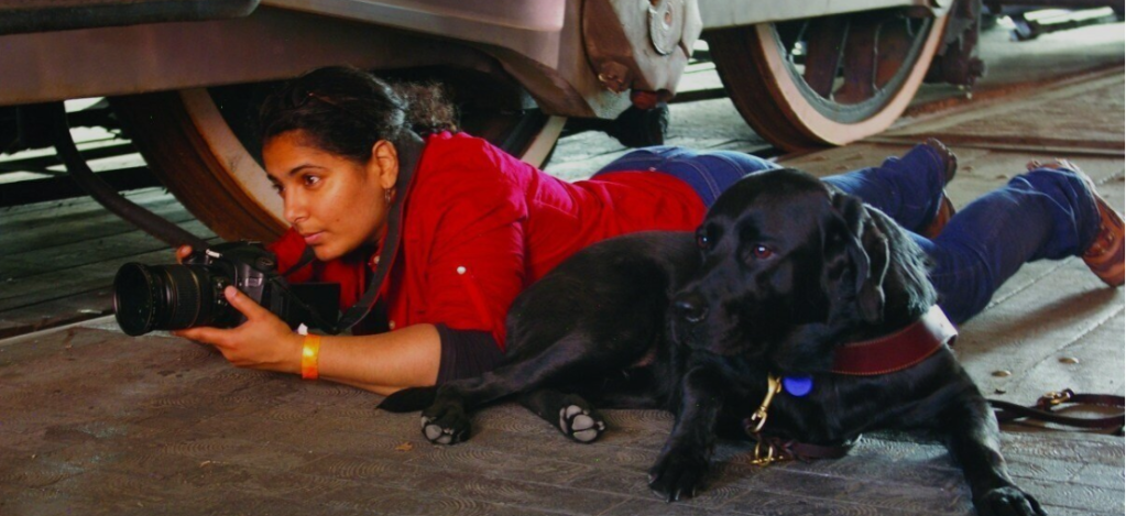 A filmmaker is lying on her stomach, propped up on her elbows, on the ground near the wheels of a non-moving train. She holds her camera, but looks over the top. She has brown skin, black hair pulled back from her face, and an expression on her face of serious concentration. She wears a short-sleeved red top, blue jeans, and a, orange and white bangle. A black labrador sits on the ground with its body up against her, gazing off camera at the same thing the filmmaker is looking at. 