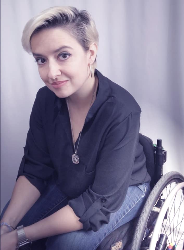 A woman with dark eyes and pale skin sits in her wheelchair. She has bleached blonde hair with dark roots cut in a short, 80s-inspired style. She wears a black top, a silver pendant, silver hoop earrings, and blue jeans.  Her hands are in her lap and her body is turned away from the camera, but her head is turned to face us. and she has a slight smile that's a bit mischievous. 