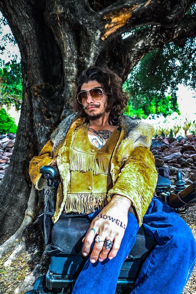 A man sits in his wheelchair outside in front of a large tree. He has long, curly drak hair, olive skin, and a beard. His mustache has been waxed to curl up at the ends. He wears large, 70s-style sunglasses, a dark yellow suede coat with a fur-lined hood and fringe at the chest and waist, and blue jeans. He has one hand on the wheelchair armrest and one on his knee.  He has tattoos on his chest and hand, and a serious expression on his face.