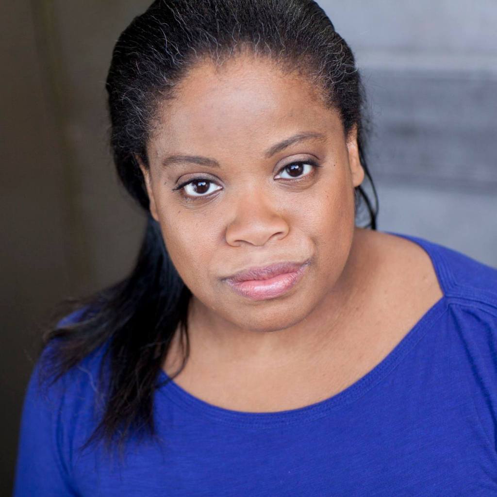 A Black woman with her hair pulled back and falling over one shoulder smiles slightly in this headshot. She wears a rich blue shirt and light pink lipstick. She leans toward the camera as if she's about to tell a fascinating story. 