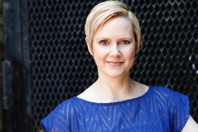 An ivory-skinned woman with short blonde hair and big hazel eyes has a perky smile in this headshot. She wears a rich blue shirt with a zig-zag pattern woven into the fabric. She stands in front of a black fence. 
