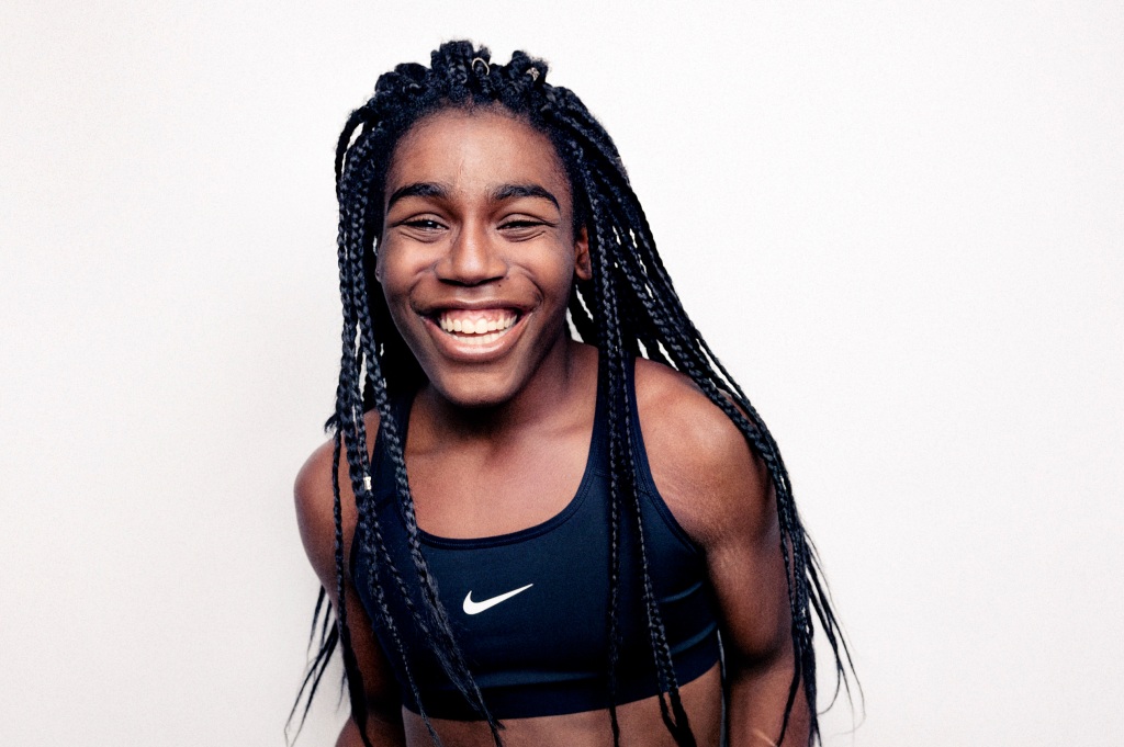A young Black girl laughs while standing against a white background. Her hair is in long, small braids that tumble down her back and over her shoulders. She wears a black Nike cropped running top.